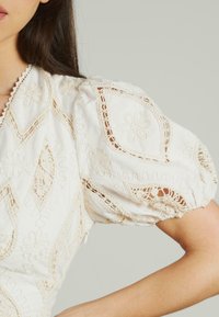 Blusa bianca ricamata con maniche a sbuffo, intricati motivi floreali e delicati profili in pizzo. Tessuto morbido con una finitura texture.