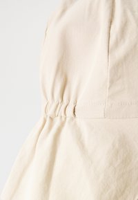 Tissu beige clair avec une surface texturée, présentant des détails froncés et un design évasé en bas. Accents cousus visibles.