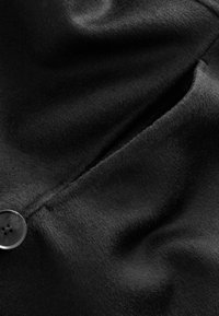 Tissu en laine noire avec une texture lisse et une légère brillance, présentant un bouton visible et des détails de couture près de l'ouverture d'une poche.