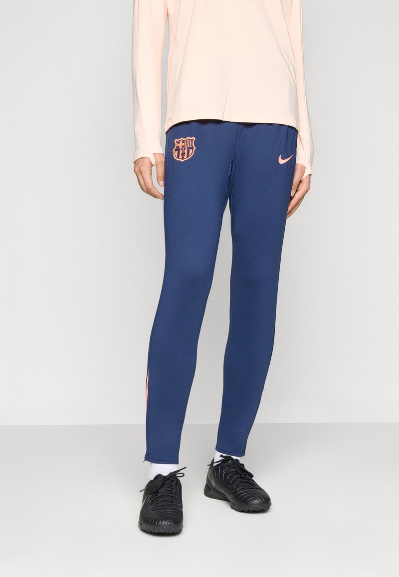 Person som har på seg marineblå Nike treningsbukser med rosa FC Barcelona og Nike logoer, svarte fotballsko og hvite sokker mot en nøytral bakgrunn.