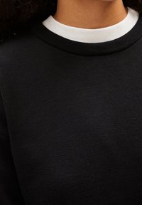 Sweat-shirt noir en tissu tricoté texturé, avec un col rond côtelé blanc. Le design est simple et minimaliste, sans ornements.