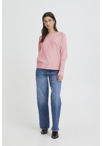 Pull en maille rose clair à coupe décontractée, col large, manches longues, associé à un jean bleu taille haute et des chaussures foncées.