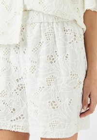 Hvit brodert skjørt med blomsterlacemønster, elastisk midje og teksturerte detaljer. Nærbilde viser det intrikate designet.