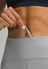 Szare legginsy sportowe z gładkiego, elastycznego materiału. Ręka poprawia jasnoszary pasek w talii, odsłaniając płaską powierzchnię skóry.