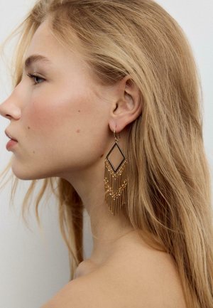 Mlajša ženska s svetlo blond lasmi nosi veliko zlato geometrično uhani z visečimi verižicami, obrnjena levo na enobarvnem ozadju.