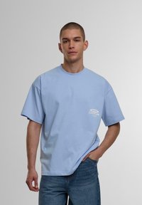 Tricou din bumbac de culoare bleu deschis, cu o croială lejeră, guler rotund și mâneci scurte. Prezintă un logo alb imprimat în zona stângă a pieptului.