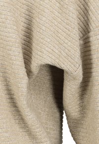 Nahaufnahme eines beigefarbenen, gerippten Strickstoffs mit weichen Falten und strukturierten, horizontalen Linien.