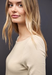 Beige, langärmliges Oberteil mit rundem Ausschnitt, aus glattem Stoff und taillierter Passform. Die Haare leicht zerzaust, mit sanften Wellen.