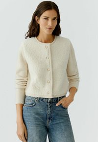 Romige textuur cardigan met ronde halslijn en zes decoratieve knopen, gecombineerd met blauwe high-waist jeans van denim. Lichte achtergrond.