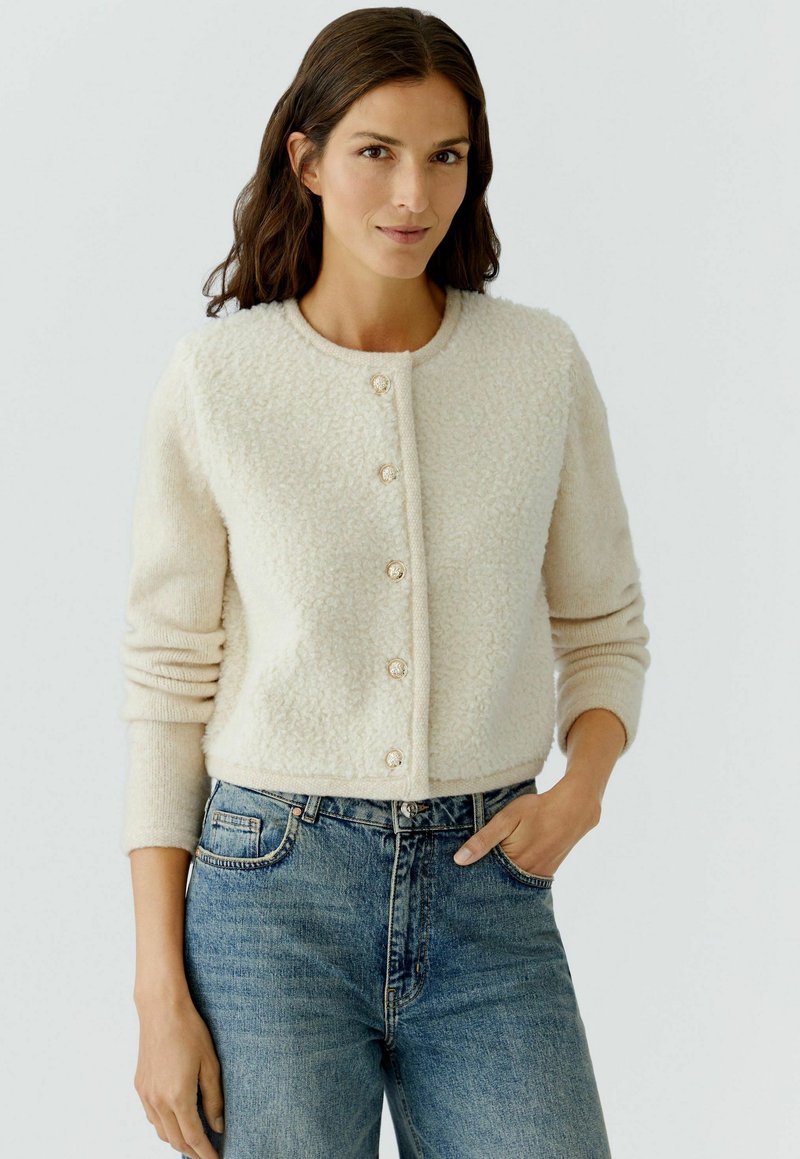Romige textuur cardigan met ronde halslijn en zes decoratieve knopen, gecombineerd met blauwe high-waist jeans van denim. Lichte achtergrond.
