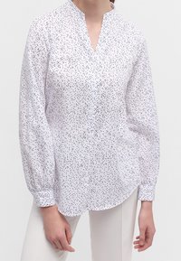 Blouse blanche à motif de petits pois noirs subtils, dotée d'un col montant et d'une fermeture à boutons, manches longues avec poignets froncés, et une coupe décontractée.
