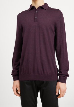 Ein Mann trägt ein dunkelviolettes Poloshirt mit langen Ärmeln und drei Knöpfen, kombiniert mit schwarzen Hosen, und steht vor einem einfarbigen weißen Hintergrund.