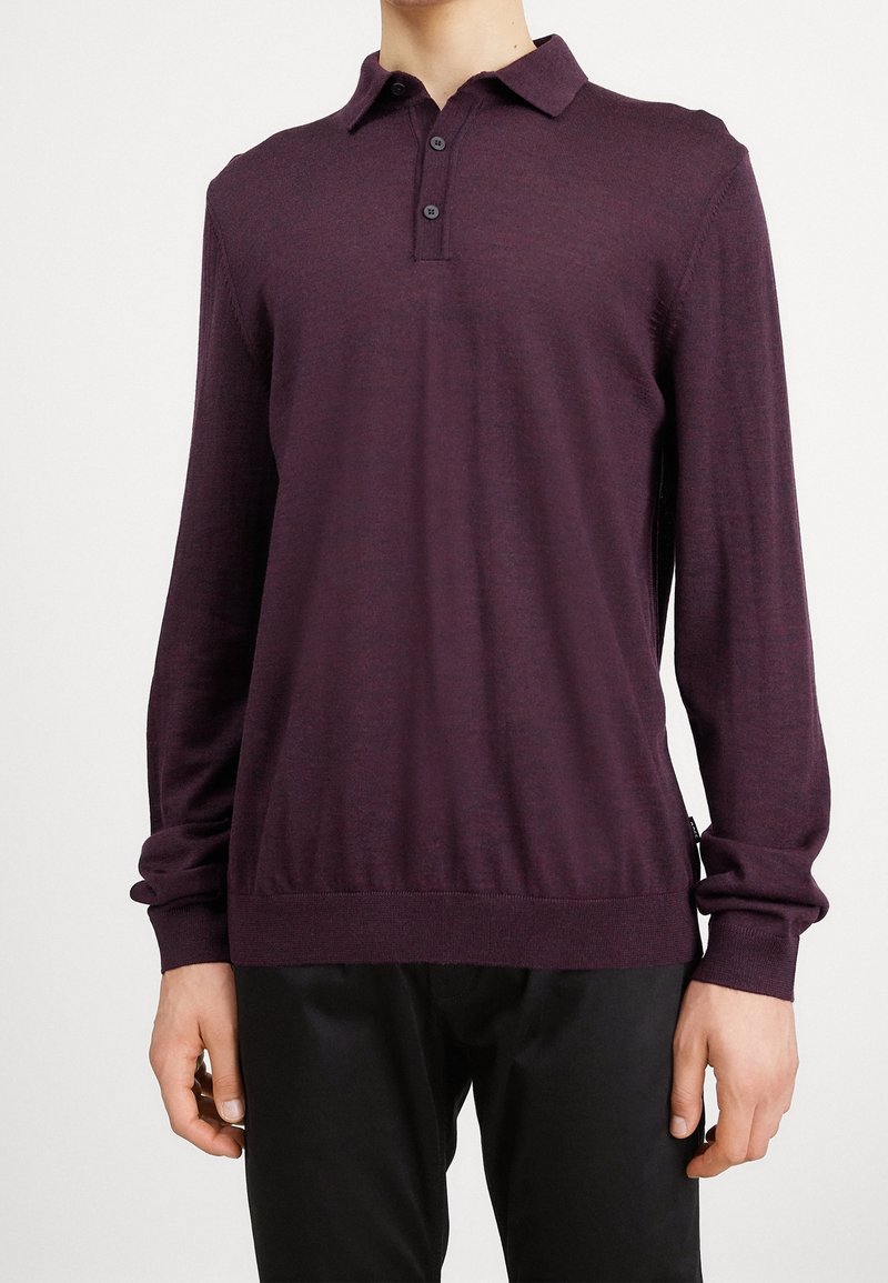 Homme portant un polo à manches longues violet foncé avec trois boutons, associé à un pantalon noir, debout devant un fond blanc uni.