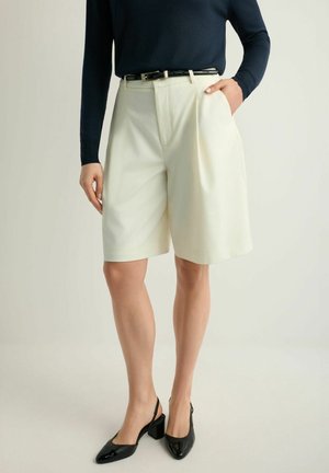 Osoba nosząca kremowe, dopasowane szorty do kolan z czarnym paskiem, granatową bluzkę z długim rękawem oraz czarne sandały na obcasie typu blok z paskiem za piętą.