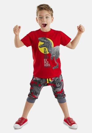 T-shirt en coton rouge avec un graphique de dinosaure et un cercle jaune. Pantalons de survêtement gris en camouflage avec des accents rouges et un cordon de serrage jaune. Baskets rouges.