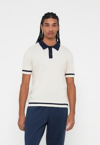 Polo majica v kremni barvi z mornarsko modrimi detajli, kratkimi rokavi, gumbasto zaponko in kontrastnimi črtami na rokavih ter robu. Mehak, strukturiran material.