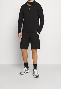 Homme portant un pull noir à zip quart sur une chemise rayée noire et jaune, un short cargo noir, des chaussettes noires et des chaussures de sport blanches.