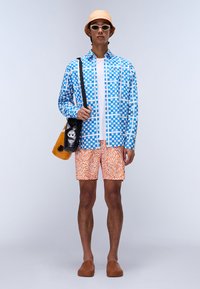 Blauw-witte geruite blouse, oranje bedrukt short, bruine instappers, beige bucket hat en witte zonnebril met een zwarte tas.