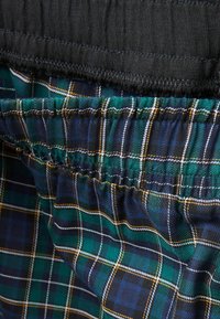 Šortky s elastickým pasem, s černou svrchní vrstvou, pod kterou se nachází modro-zelený kostkovaný vzor s bílými a žlutými pruhy.