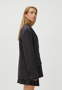 Blazer preto de carvão escuro com um corte ajustado, design de um só botão e dois botões, combinado com uma saia preta plissada. Textura suave.