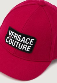 Sarkans vilnas beisbola cepure ar melnu izšūtu uzlīmi, kurā rakstīts "VERSACE JEANS COUTURE" baltā tekstā. Strukturēta dizaina ar izšūšanas detaļām.
