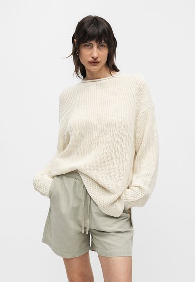 Γυναίκα που φοράει ένα φαρδύ κρεμ πλεκτό πουλόβερ και ανοιχτό γκρι σορτς με κορδόνι, στέκεται με τα χέρια στις τσέπες μπροστά σε απλό λευκό φόντο.