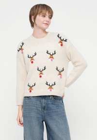 Sieviete, kura valkā bēšas krāsas džemperi ar vairākiem ziemeļbriežu sejas attēliem, kuriem ir sarkanas un rozā pom-pom deguni, apvienojot to ar zilo džinsu biksēm, uz baltas fona.