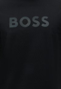 Μαύρο ύφασμα με τη λέξη "BOSS" τυπωμένη με μεγάλα, έντονα, σκούρα γκρί γράμματα στο κέντρο.