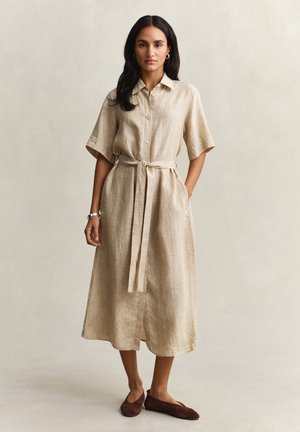 Vrouw met lang donker haar, gekleed in een beige midi-jurk met knopen en een gestrikte tailleband, en bruine platte schoenen, staand tegen een effen lichte achtergrond.