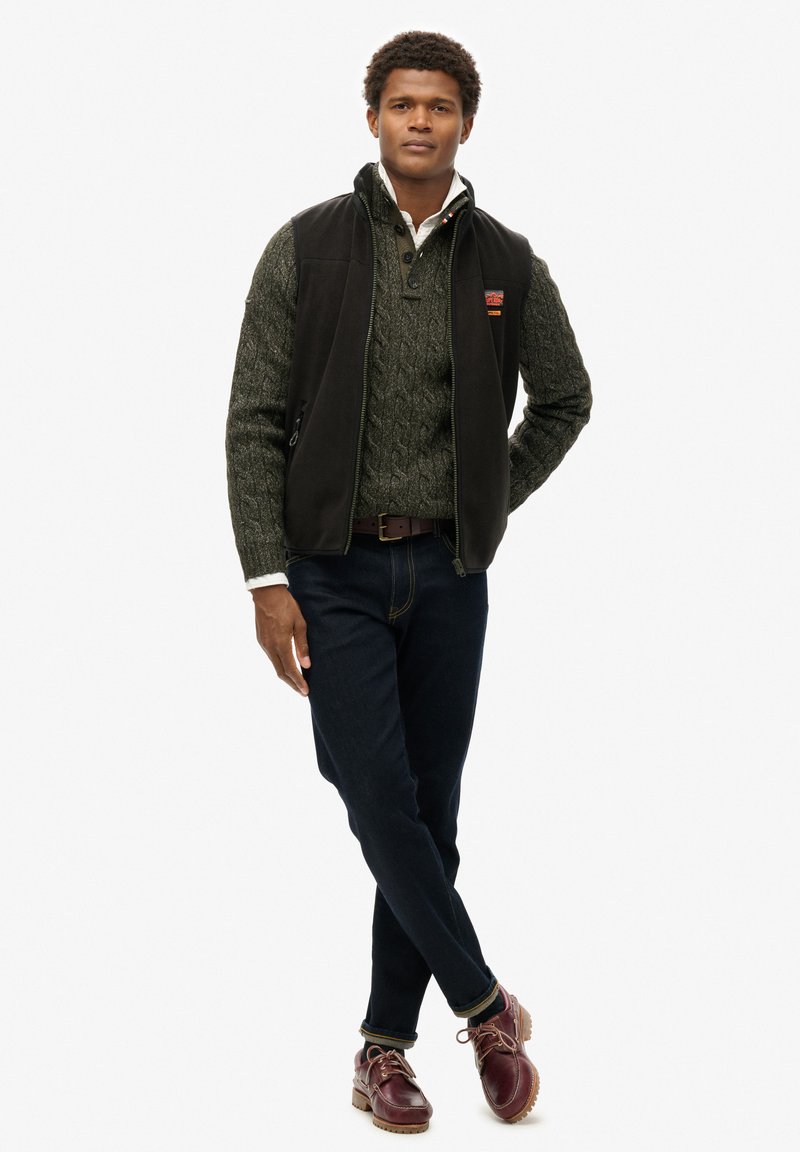 Gilet en polaire noir sur un pull en tricot torsadé vert, avec un jean bleu marine et des chaussures à lacets bordeaux. Détails remarquables : un patch avec logo sur le gilet.