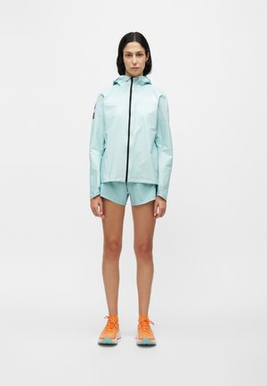 Kvinde iført en lyseblå hættetrøje og shorts med lyse orange sneakers står mod en ensfarvet hvid baggrund.