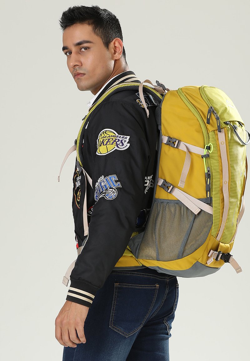 Zaino giallo e grigio con più scomparti, tasche laterali in rete e spallacci regolabili, indossato con una giacca bomber nera con toppe.