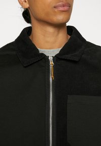 Veste zippée noire avec un col en velours côtelé contrastant, une poche à rabat et un tissu lisse, avec un détail de tirette de fermeture éclair orange.