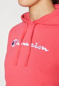 Felpa corallo con cappuccio e cordino, dotata del logo "Champion" ricamato in bianco. Materiale morbido in misto cotone, dettagli di cucitura visibili.