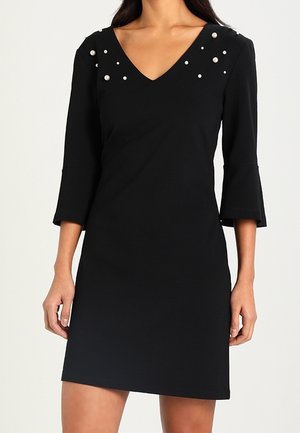 Robe noire avec un décolleté en V et des manches trois-quarts, ornée de détails en forme de perles aux épaules. Tissu lisse, silhouette ajustée.