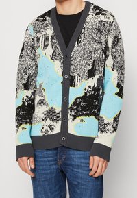 Cardigan en tricot avec un motif abstrait multicolore dans des nuances de bleu, noir et crème. Comprend une fermeture à bouton et des poignets côtelés.