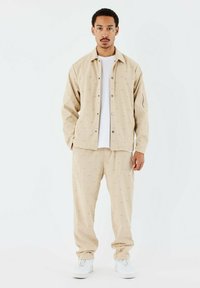 Conjunto de chaqueta y pantalones en beige a juego, con un sutil patrón texturizado de logo. Camiseta blanca debajo, combinada con zapatillas blancas.