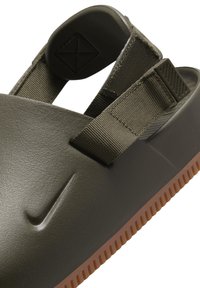 Olivinės spalvos šlepetės su tekstūruotu viršumi, reguliuojama juosta ir rudomis gumos padais, turinčiais gumbelius sukibimui. Įspaustas Nike logotipas.