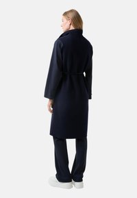 Navy wool-blend long coat with a high collar, cinched waist, and buttoned sleeves. Worn over matching trousers and white sneakers.