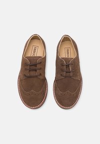 Ruskeat mokkaiset brogue-kengät siipikuvioinnilla, joissa on reikäkuviointia, litteät nauhat ja kontrastinen valkoinen pohja.