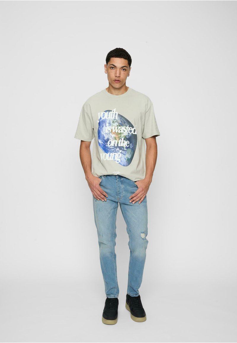 T-shirt gris clair avec impression graphique représentant la Terre et du texte, associée à un jean bleu délavé et des chaussures noires avec une semelle texturée.