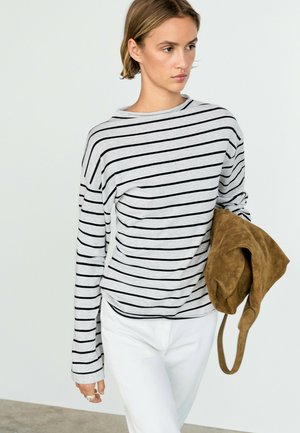 Femme portant une chemise grise à manches longues avec des rayures horizontales noires et un pantalon blanc, tenant un sac en daim marron dans sa main gauche.