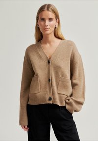 Beige kötött kardigán V-nyakkal, hosszú ujjakkal, két elülső zsebbel, bordás szegéllyel és fekete gombokkal a záráshoz. Puha tapintású.