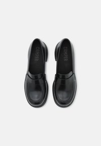 Chaussures en cuir noir avec un bout arrondi, un talon plat et des détails de couture sur le dessus. La semelle intérieure porte le nom de la marque "CAMPER."
