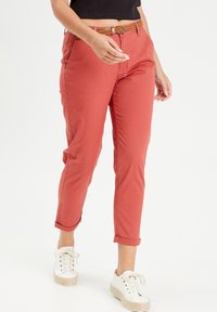 Pantalones de color óxido, ajustados y con puños enrollados, que presentan un cinturón trenzado color camel, hechos de tela ligera, estilizados con zapatillas blancas.