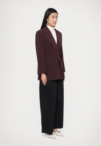 Bordó színű, övvel megkötött körbepántos blézer, széles ujjakkal és oldalzsebekkel, fekete bő szárú nadrággal és fehér, hegyes orrú cipővel párosítva.