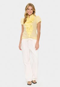 Blusa gialla con dettagli arricciati e maniche corte, abbinata a pantaloni bianchi a gamba larga. Il modello sorride su uno sfondo grigio chiaro.