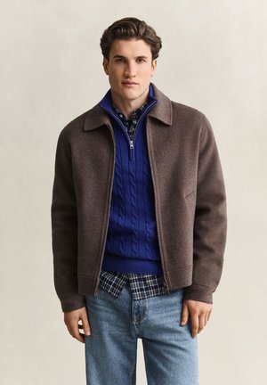 Junger Mann trägt eine braune Jacke über einem blauen Zopfmuster-Pullover und einem karierten Hemd, kombiniert mit hellblauen Jeans, stehend vor einem neutralen Hintergrund.