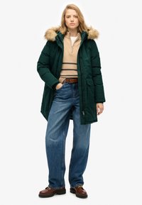Chaqueta acolchada verde con capucha forrada de pelo, combinada con un suéter a rayas beige y jeans anchos azules. Zapatos marrones completan el conjunto.
