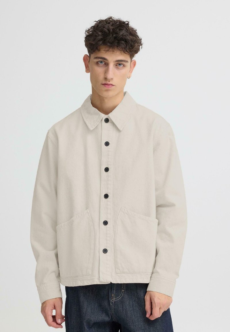 Veste beige clair avec col, fermeture à boutons sur le devant, deux grandes poches plaquées, et boutons noirs. Confectionnée dans un tissu robuste.